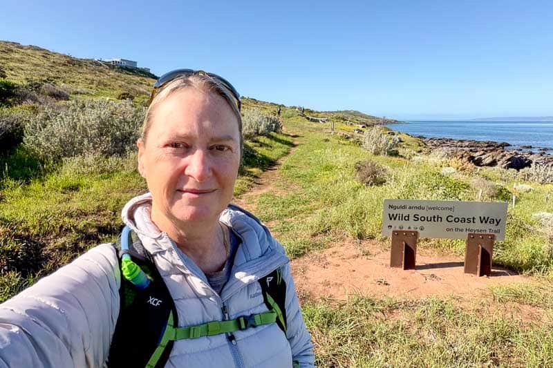 Sixty before 60 - the New & Improved Bucket List 7 Josie taking a selfie on a coastal footpath with greenery and rocks. A sign nearby reads “Wild South Coast Way on the Heysen Trail.” Blue sky and sea visible in the background.