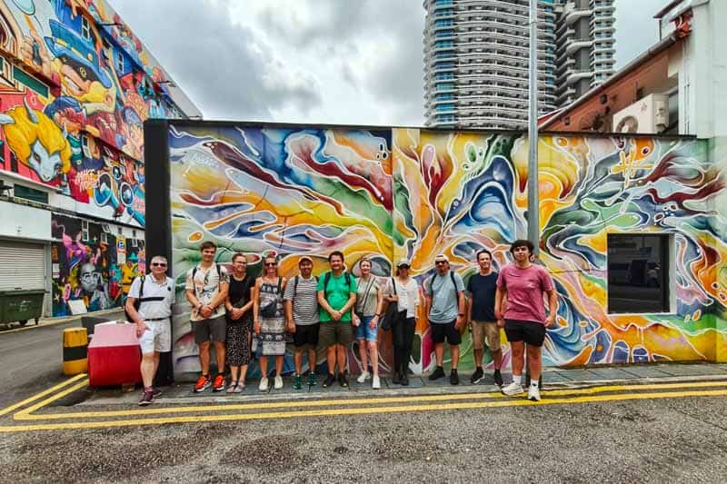 32 Fun Things to do in Singapore on a Budget 8 A group of twelve people stands in front of a colourful graffiti mural in an urban area, with tall buildings and other vibrant street art in the background. The group poses casually on a paved street.