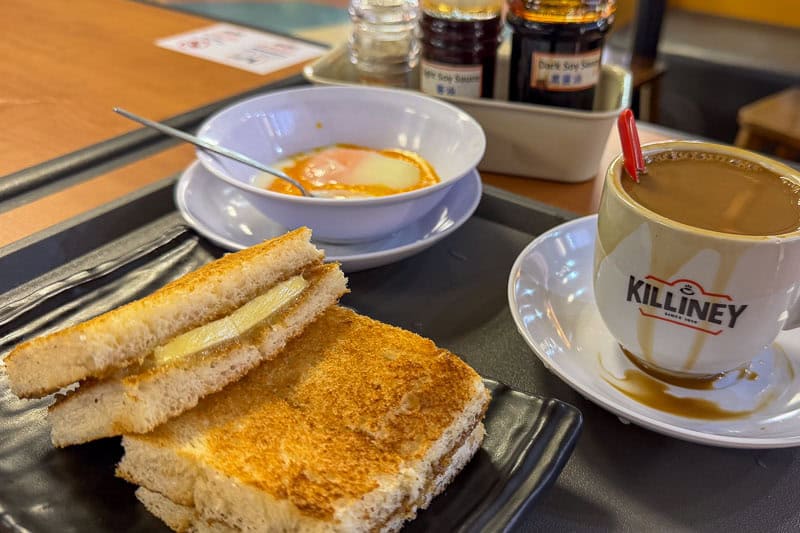 Where to Eat Kaya Toast in Singapore: 9 Places I've Tried 22 A breakfast set with buttered toast, soft-boiled eggs in a bowl, and a cup of coffee on a tray. Soy sauce and condiments are in the background on the table. The cup is branded with the word “KILLINEY”.