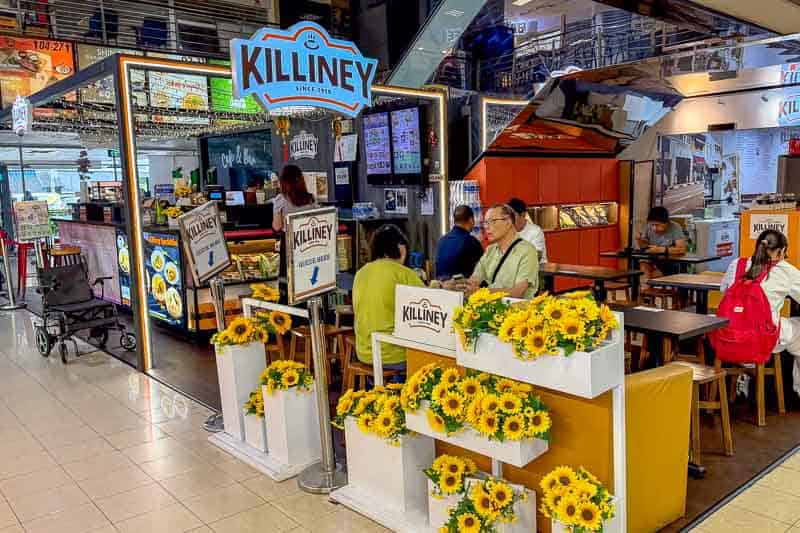 Where to Eat Kaya Toast in Singapore: 9 Places I've Tried 21 A food stall named Killiney, with several people seated at tables. The area is decorated with bright yellow artificial sunflowers, and menus are visible on the tables and displays behind the counter.