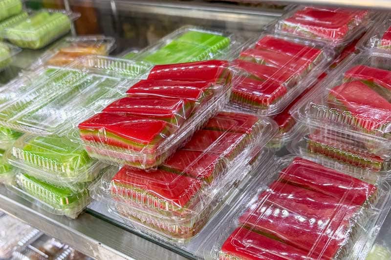 Must-Try Food in Singapore: What to Eat on Your Visit 17 Stacks of clear plastic containers hold green and red layered puddings, neatly arranged on metal shelves in what appears to be a refrigerated display cabinet at a shop or market.