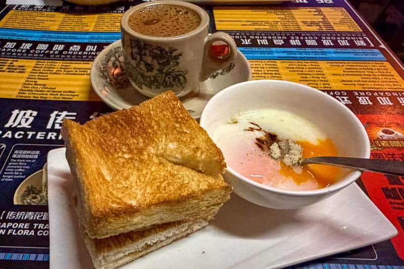 Where to Eat Kaya Toast in Singapore: 9 Places I've Tried 20 Two thick slices of toasted bread, a bowl with two soft-boiled eggs and a spoon, and a cup of coffee on a saucer sit on a colourful menu-covered table.