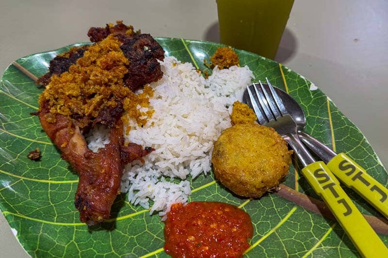 Must-Try Food in Singapore: What to Eat on Your Visit 7 A plate with white rice, fried chicken, a round fried patty, red chilli sauce, and yellow crumbs, served on a leaf-patterned plate with a fork, spoon, and a yellow cup in the background.