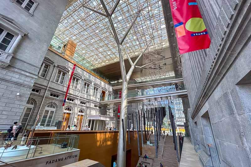 32 Fun Things to do in Singapore on a Budget 14 Spacious atrium with a glass ceiling, metal tree-like columns, and red banners. People walk around modern architectural features, with historic building façades visible alongside contemporary structures.