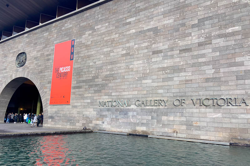 30 Free Things to Do in Melbourne 4 Large stone wall of the National Gallery of Victoria with water in front, a group of people near the arched entrance, and a red banner reading “The Picasso Century” displayed on the wall.