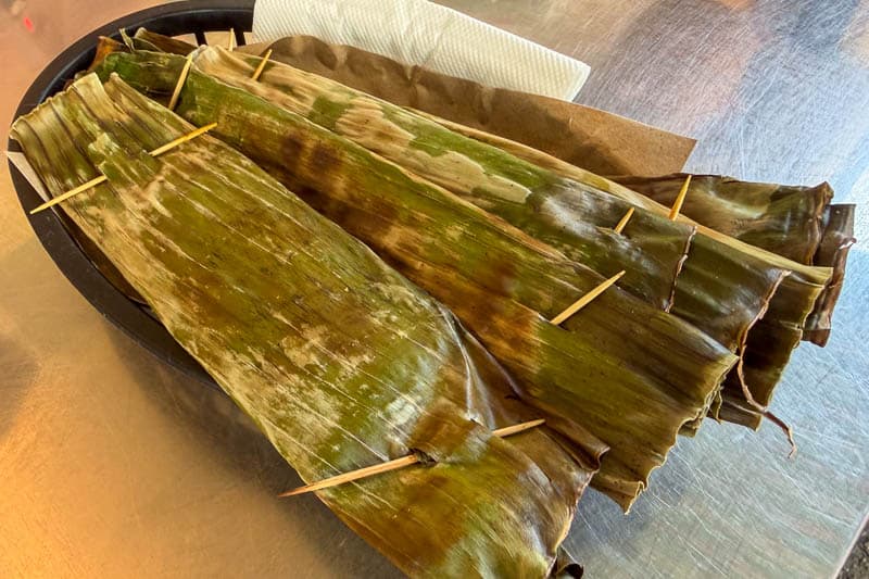 Must-Try Food in Singapore: What to Eat on Your Visit 19 Three tamales wrapped in green banana leaves, secured with cocktail sticks, rest on a black plate next to a brown paper serviette and a white paper towel on a metallic surface.