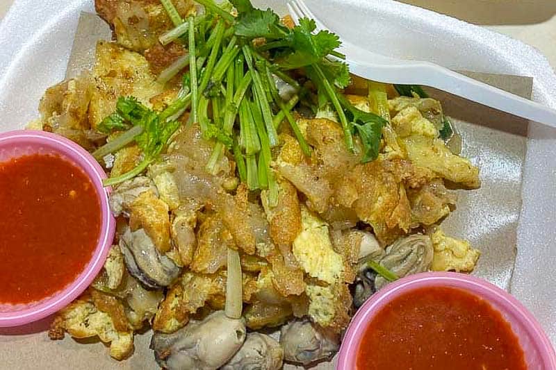 Must-Try Food in Singapore: What to Eat on Your Visit 14 A serving of fried oyster omelette topped with fresh coriander, accompanied by two portions of red dipping sauce in pink plastic pots, served on a disposable plate with a white plastic fork.