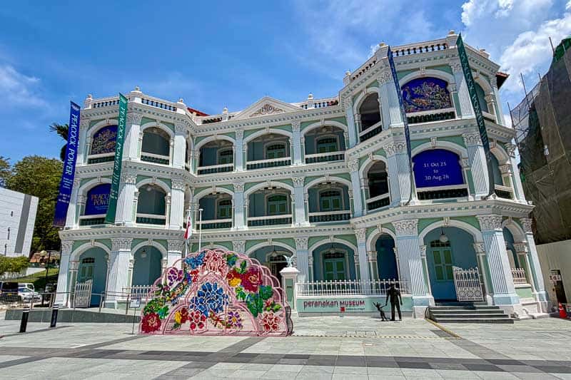 32 Fun Things to do in Singapore on a Budget 12 A three-storey colonial-style building with ornate white and blue trim, banners, and floral decorations out front. Signs indicate it is the Peranakan Museum. The sky is clear and sunny.
