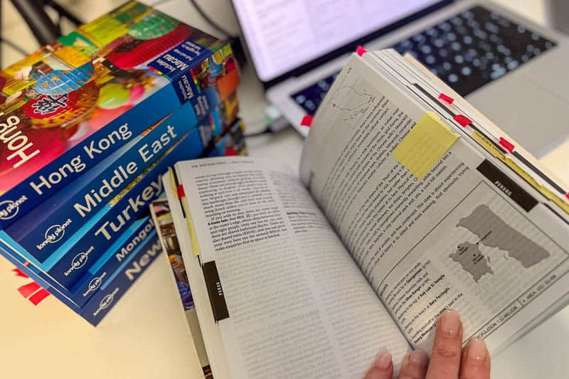 Do You Still Need Travel Guidebooks? 5 A hand holds open a travel guidebook with text and a map, next to a stack of Lonely Planet guidebooks for Hong Kong, Middle East, Turkey, and New York, with a laptop in the background.