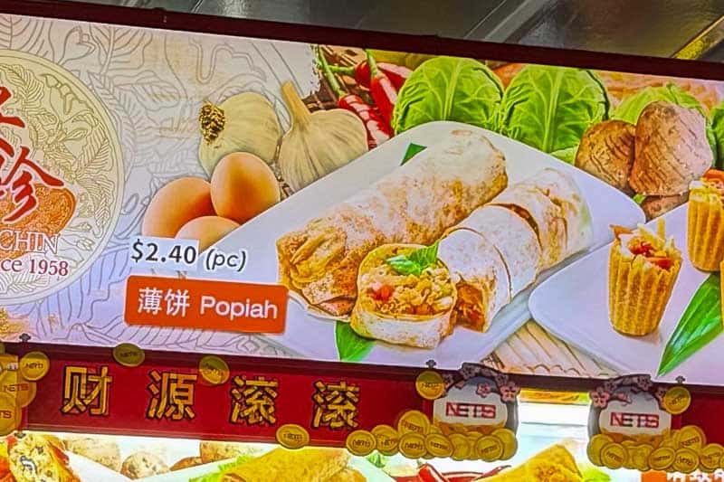 Must-Try Food in Singapore: What to Eat on Your Visit 11 A food stall sign shows a dish called “Popiah” priced at £2.40 per piece. The image features sliced popiah rolls filled with vegetables, garlic, and lettuce, along with decorative ingredients in the background.