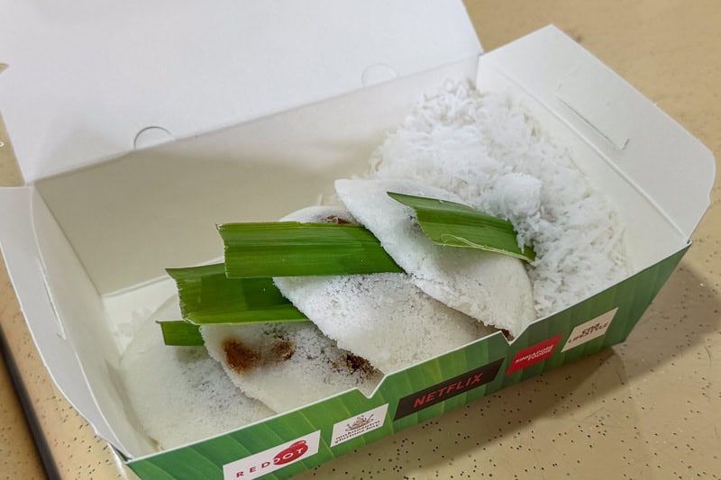 Must-Try Food in Singapore: What to Eat on Your Visit 16 A takeaway box containing several white steamed rice cakes topped with grated coconut, with green pandan leaves placed on top. The box has a green exterior with Netflix and other branding.