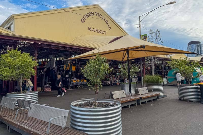 30 Free Things to Do in Melbourne 8 The image shows the entrance to Queen Victoria Market with outdoor seating, trees in large metal planters, and people walking past stalls selling various goods under yellow canopies. The market building is pale yellow with a sign visible.