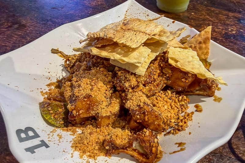 Must-Try Food in Singapore: What to Eat on Your Visit 12 A plate of chicken pieces coated in a dark sauce, topped with crushed peanuts and crispy flatbread pieces, served on a white, irregular-shaped dish labelled “1B.”.
