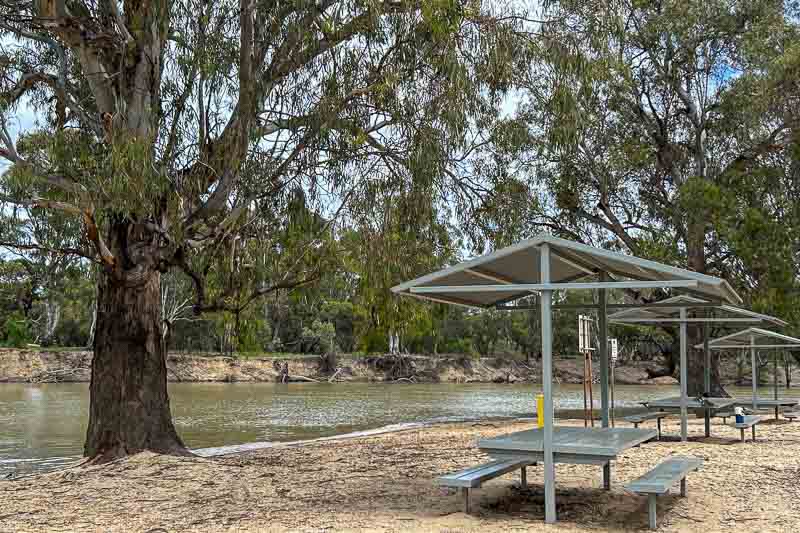 13 Things to Do in Hay, NSW 10 Picnic tables with metal shelters are set on sandy ground by a river. Large trees line the riverbank, and the water appears calm. The scene is outdoors with natural greenery and an overcast sky.