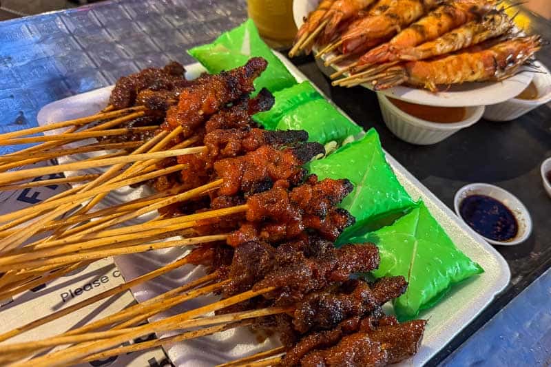 Must-Try Food in Singapore: What to Eat on Your Visit 6 A tray of grilled meat skewers, bright green packets of rice, and grilled prawns on skewers. Two sauces in small bowls are visible on a table with a clear plastic cover.