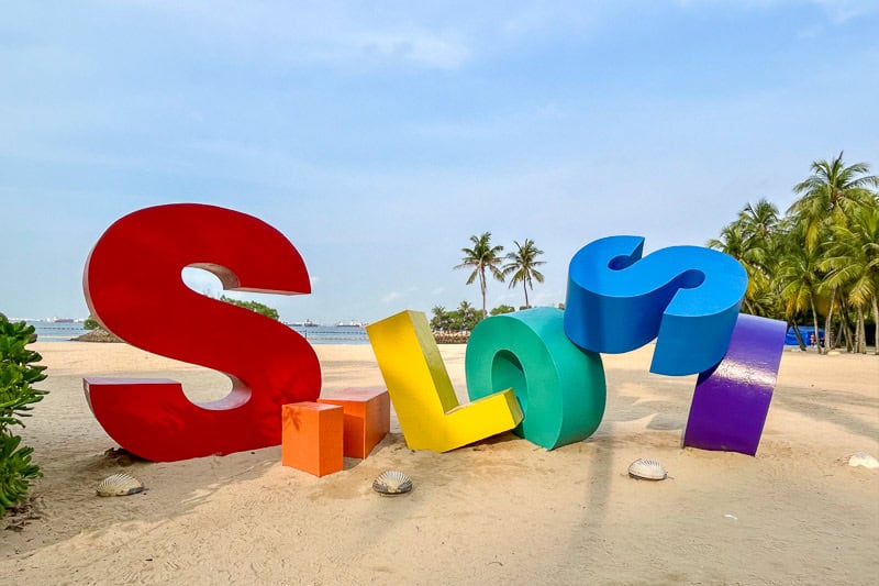 32 Fun Things to do in Singapore on a Budget 21 Large, colourful 3D letters spell “Sentosa” on a sandy beach with palm trees and blue sky in the background. The letters are red, orange, yellow, green, blue, and purple.