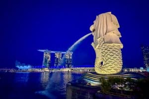 32 Fun Things to do in Singapore on a Budget 49 The Merlion statue in Singapore spouts water into Marina Bay at night. The illuminated Marina Bay Sands hotel and ArtScience Museum are visible in the background against a deep blue sky.
