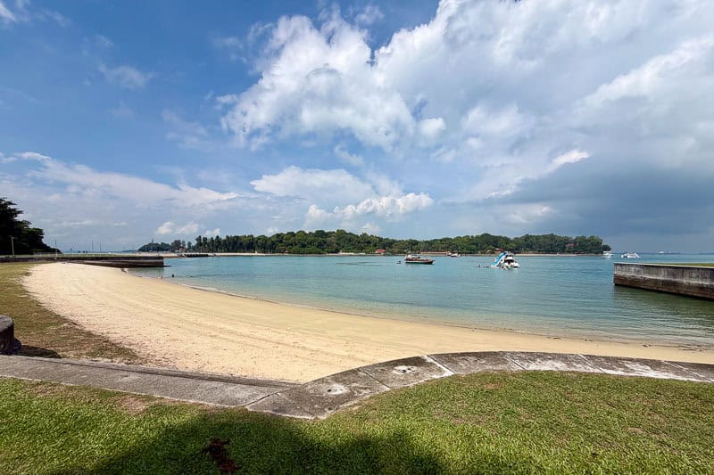 32 Fun Things to do in Singapore on a Budget 26 A sandy beach curves along clear, calm water with a grassy area in the foreground. Several small boats are on the water, and an island covered in green trees is visible in the distance under a partly cloudy sky.