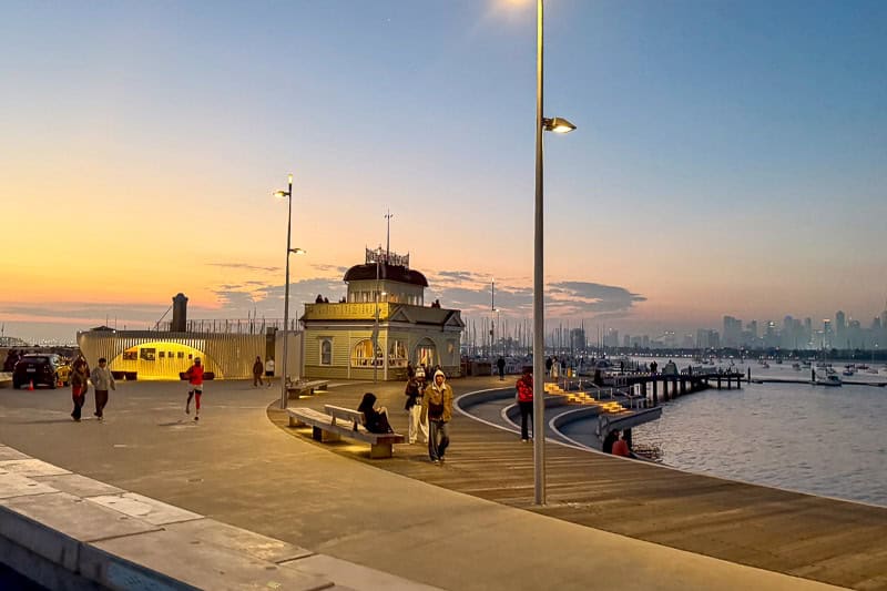 30 Free Things to Do in Melbourne 15 People walk and sit near a promenade by the water at sunset. A historic building and street lamps are visible, with boats and a city skyline in the background. The sky is clear with soft orange and blue hues.