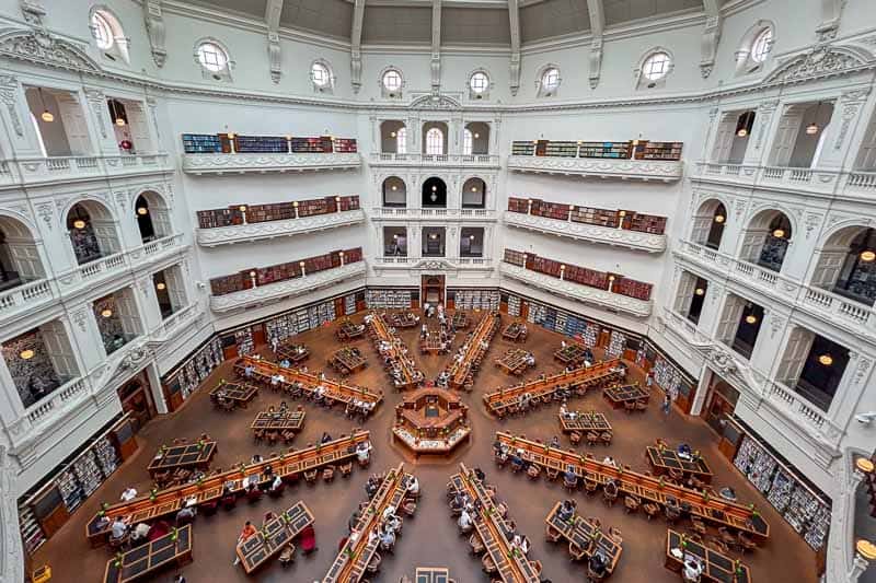 30 Free Things to Do in Melbourne 5 A large, circular library reading room with multiple floors and white walls. People sit at wooden tables working or reading. Bookshelves line the walls on every level, and a domed ceiling is visible at the top.