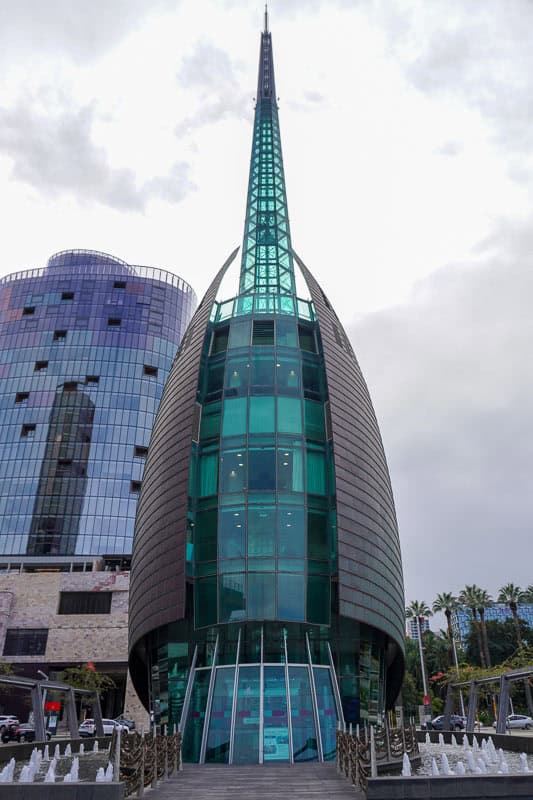 Essential Things to do in Perth on Your First Visit 7 A modern glass and steel building with a pointed, sail-like shape stands in front of a taller curved building. Palm trees and a cloudy sky are visible in the background.