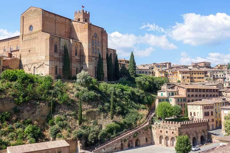 20 Fun Things to Do in Siena, Italy (Plus Two Extras!) 4 A large, red-brick gothic church stands atop a green hillside overlooking an Italian city with terracotta-roofed buildings.