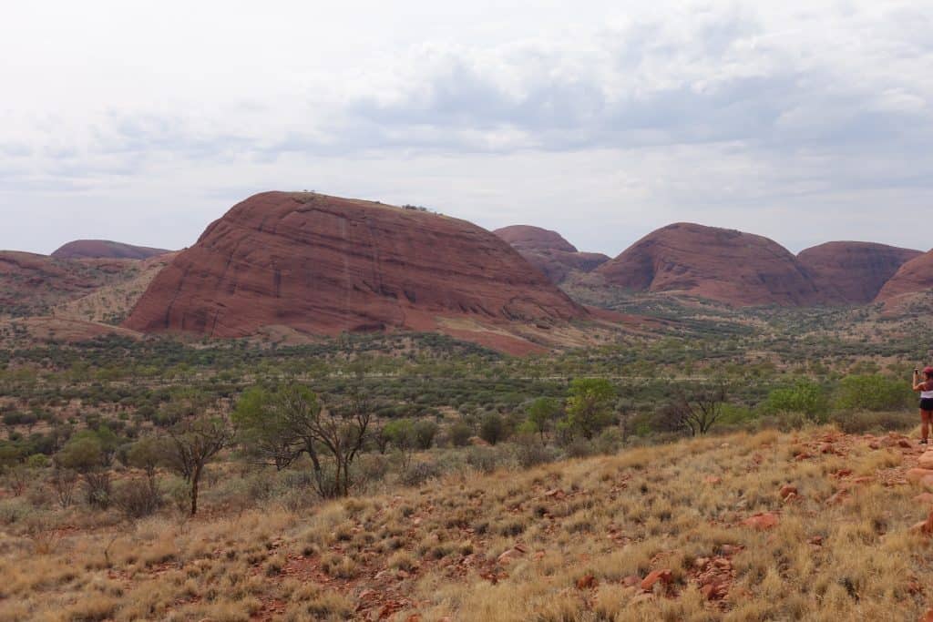 Alice Springs to Uluru on a Budget - Visit the Red Centre 7 Large red rock formations rise above a dry, scrubby landscape under a cloudy sky.